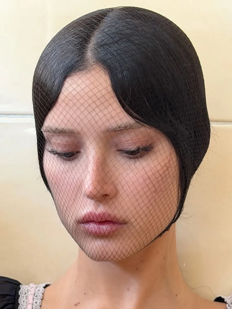 Fine black fishnet veil worn over a face with dark hair, against cream tiles